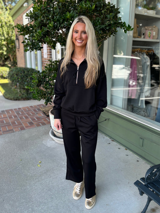 Woman in black outfit standing on a sidewalk with greenery and a building in the background