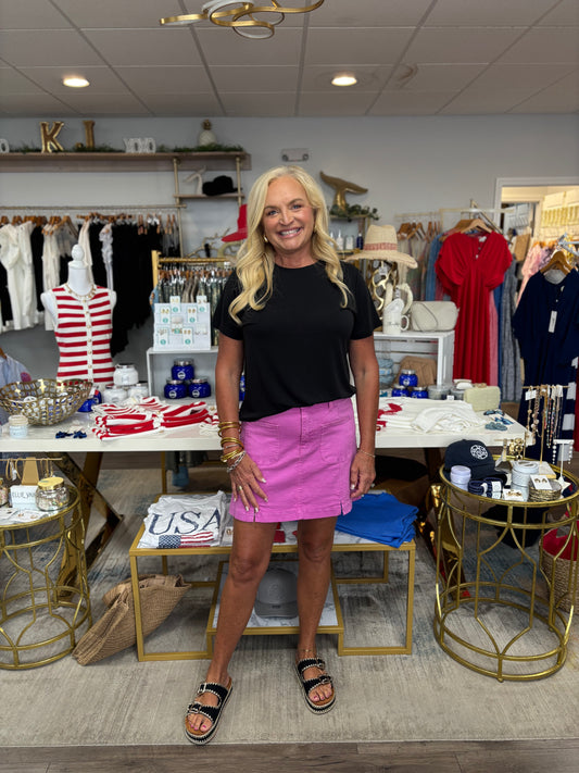 Woman standing in a clothing store wearing a black top and pink skirt.