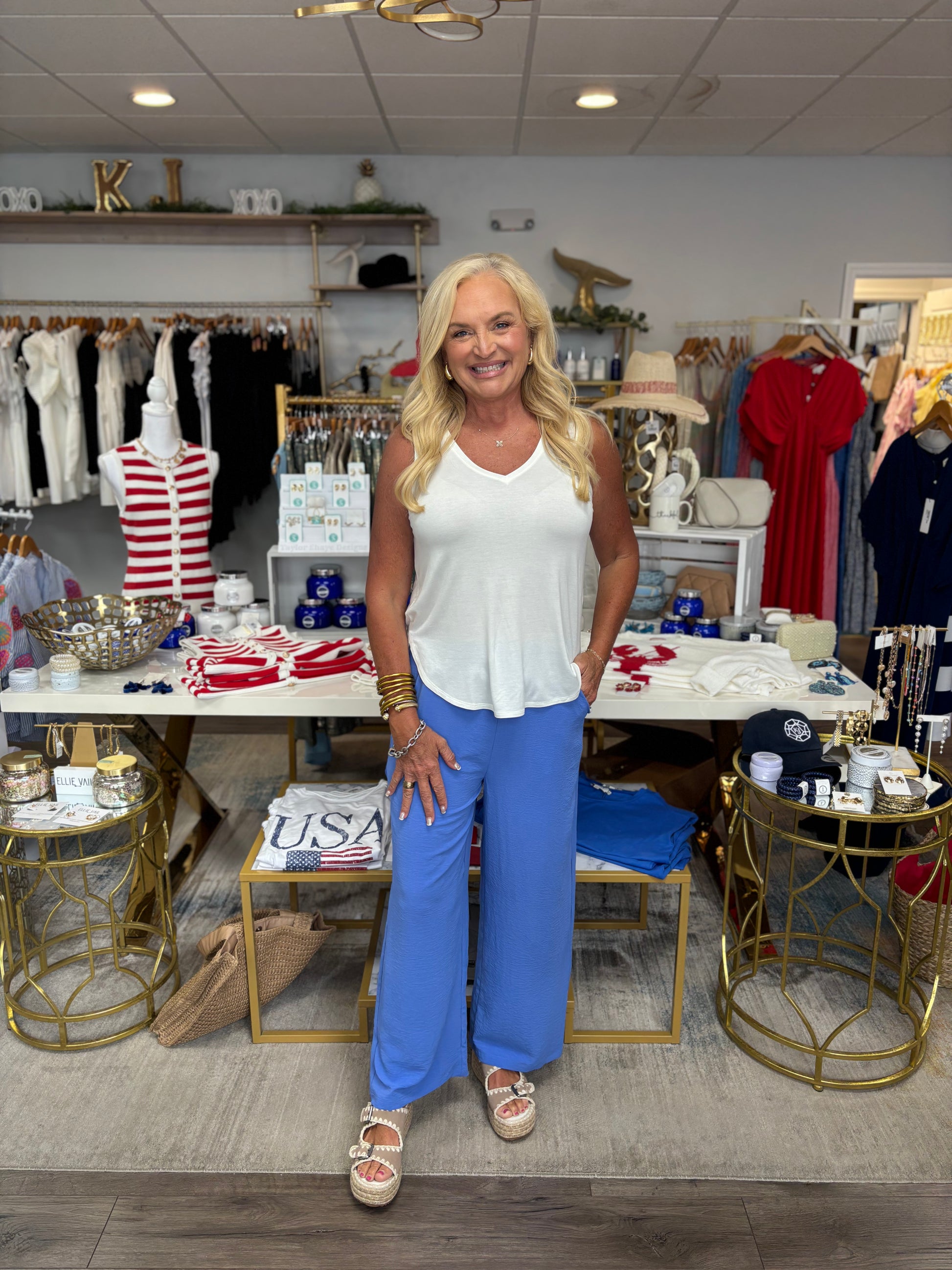 Woman in a store wearing a white top and blue pants, surrounded by clothing and decor.