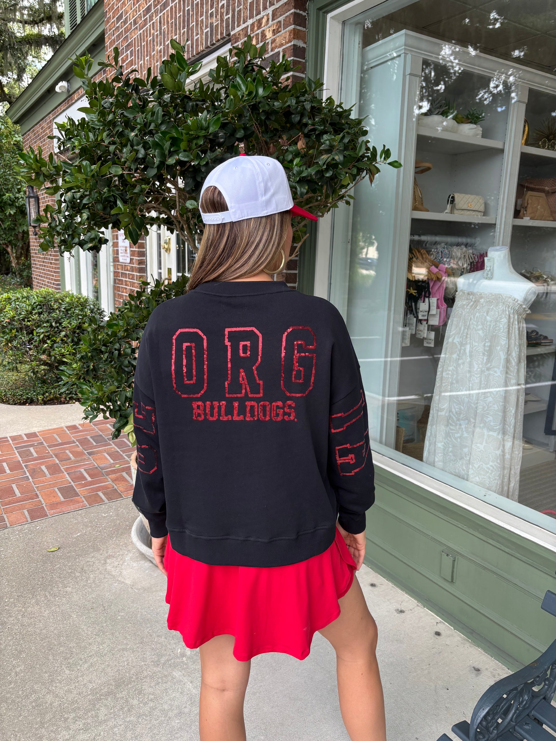 Person wearing a black sweatshirt with 'ORG Bulldogs' text, red skirt, and white cap in front of a store window.