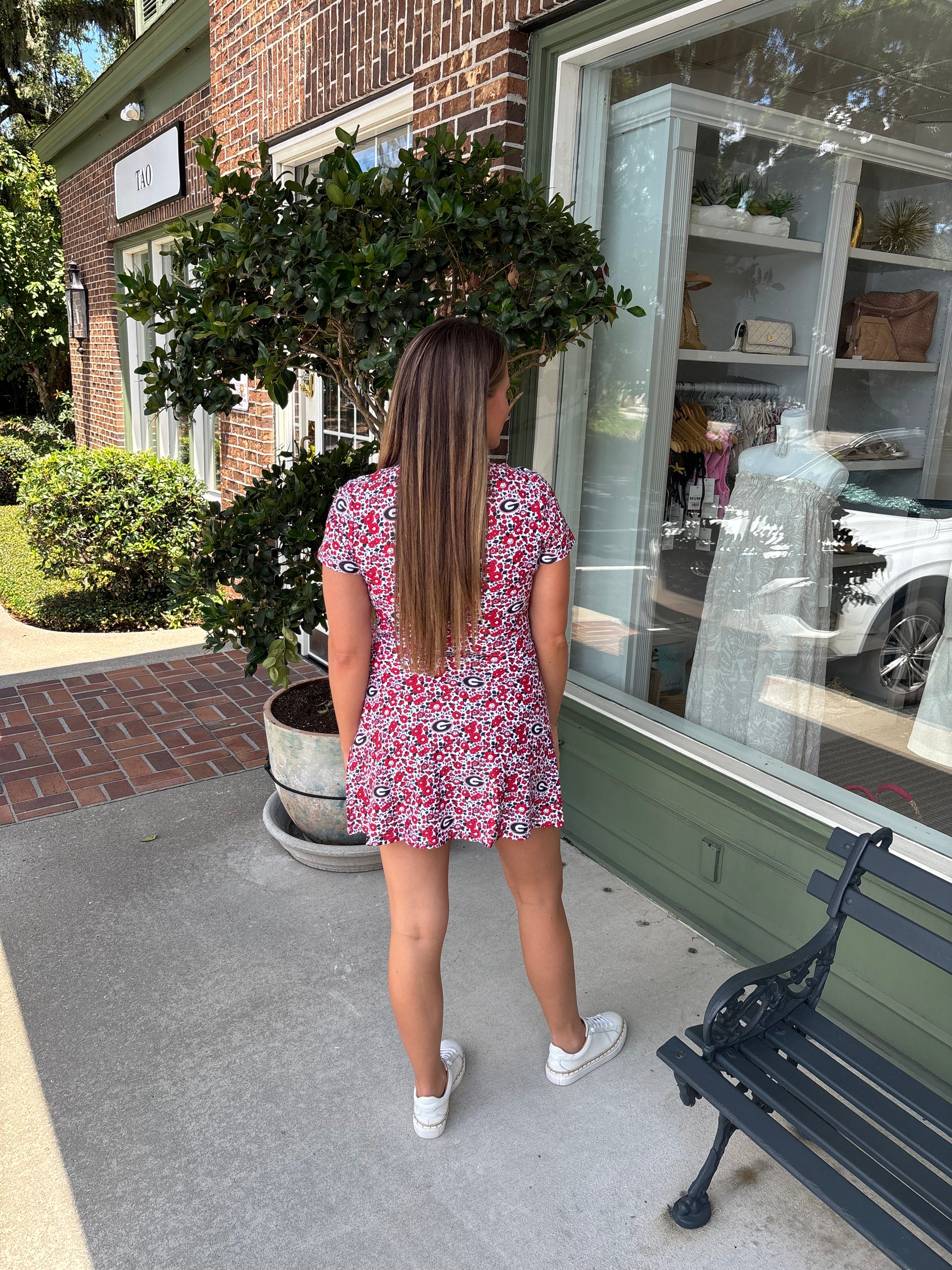 Woman in a red floral dress standing outside a store with a bench and potted plant in the foreground.