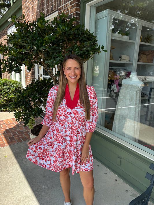 Woman in a red floral dress standing outside a store with a brick building and greenery in the background.