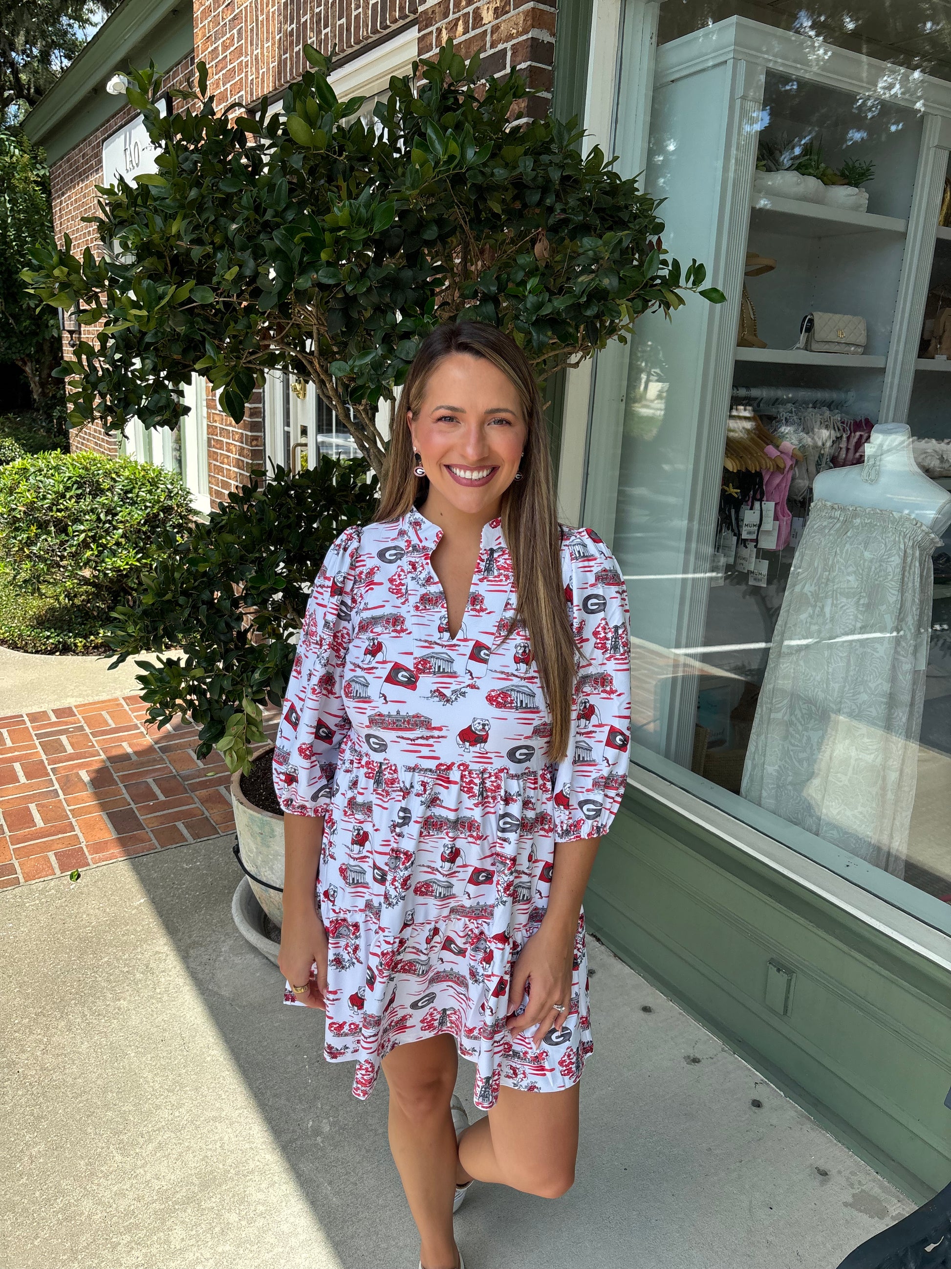 Woman in a patterned dress standing outside a building with a plant and window in the background.