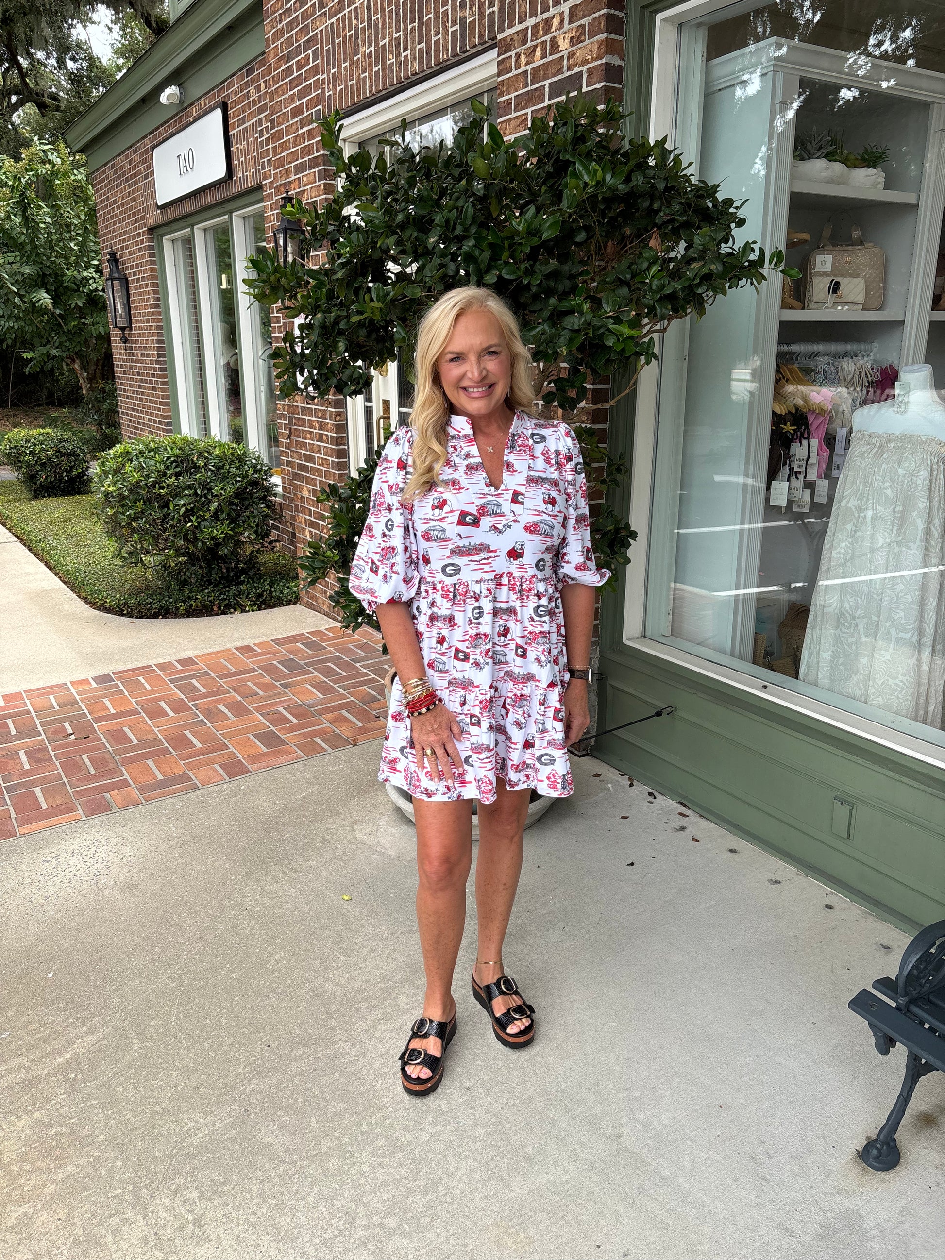 Woman in a patterned dress standing outside a store with brick walls and greenery.