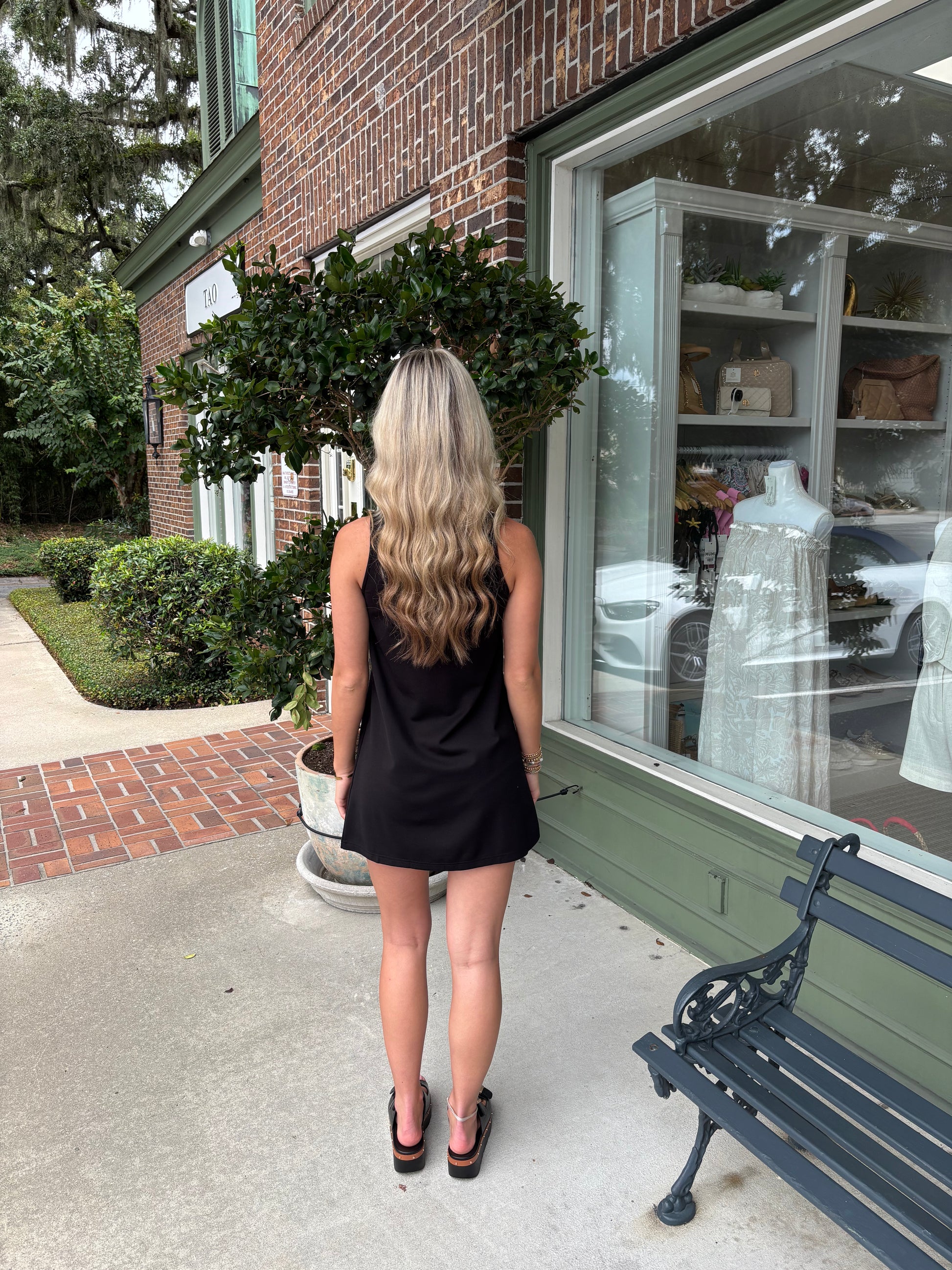 Woman in a black dress standing on a sidewalk next to a store window.