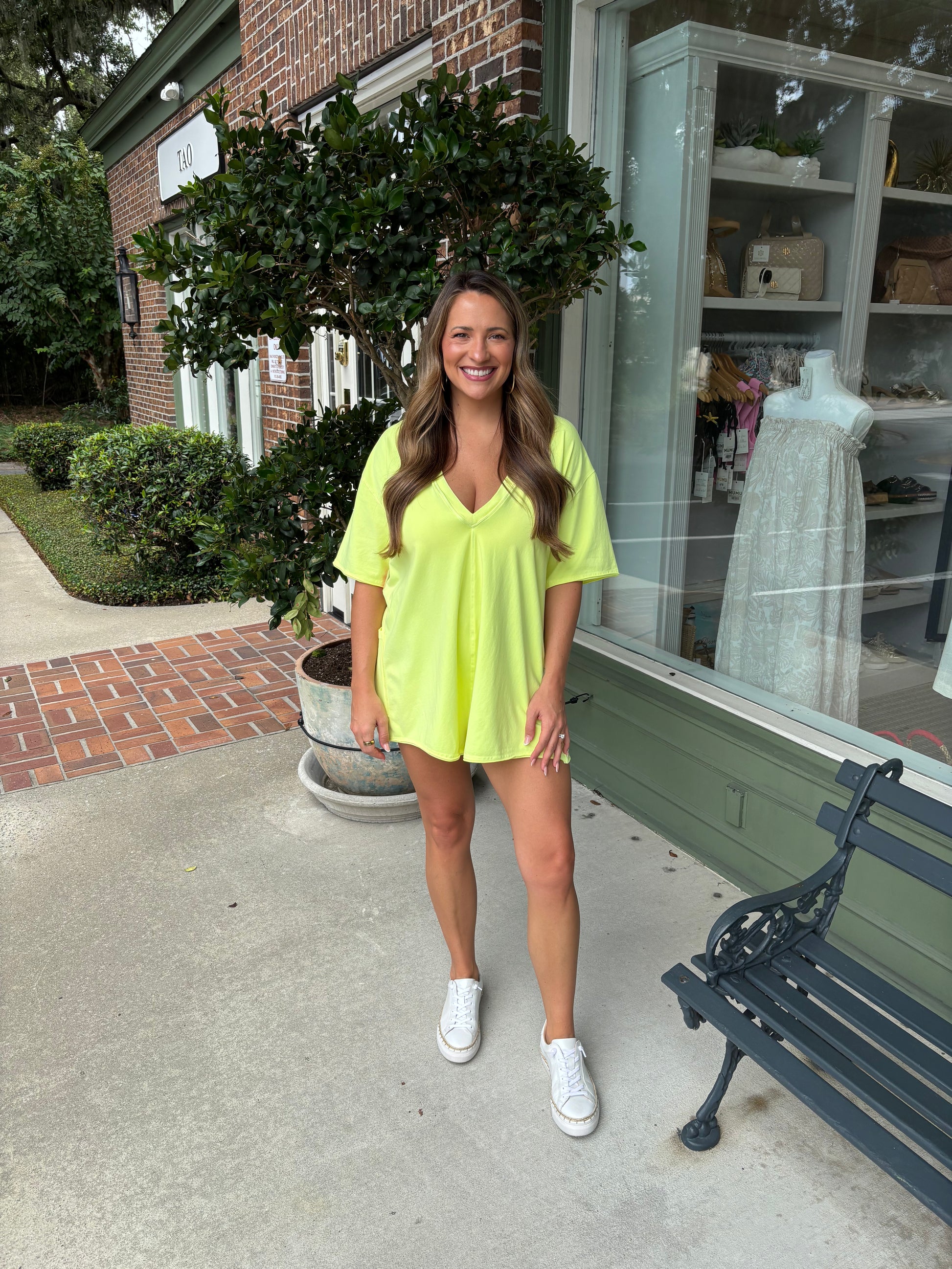 Woman in a bright yellow outfit standing on a sidewalk with a building and bench in the background.