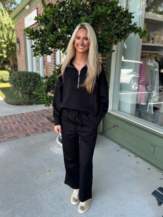 Woman in a black outfit standing on a sidewalk with a store and greenery in the background