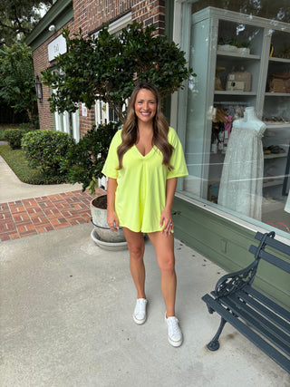 Woman in a bright yellow outfit standing on a sidewalk with a building and bench in the background.