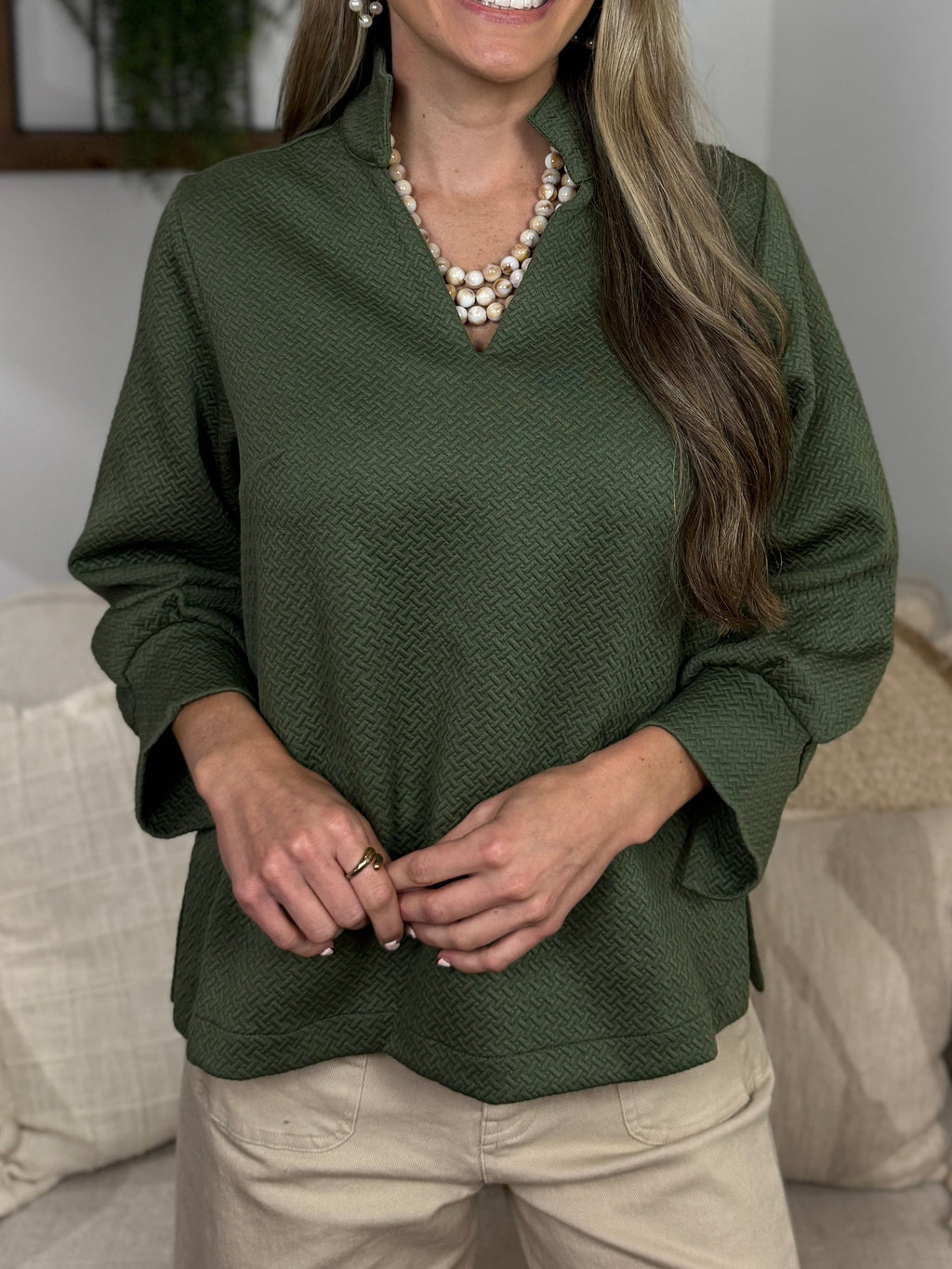 A woman with blonde hair stands in a living room setting wearing a textured olive green long-sleeve blouse and beige pants. She is smiling and posing in front of a light-colored sofa with neutral pillows.