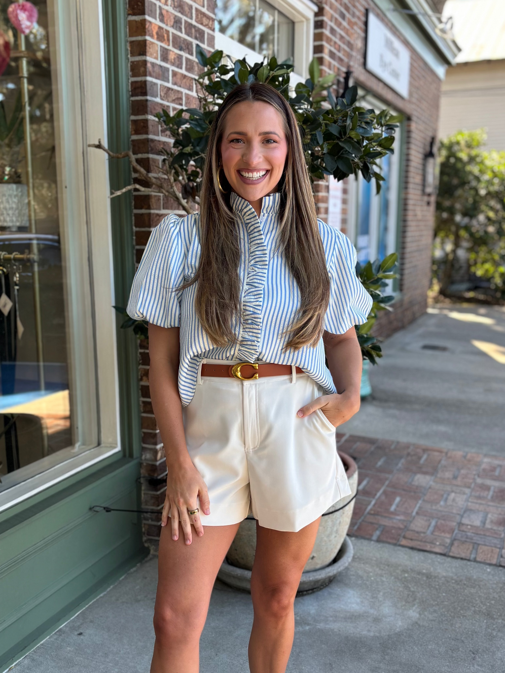 Woman standing on a sidewalk in front of a store window, wearing a striped shirt and white shorts.
