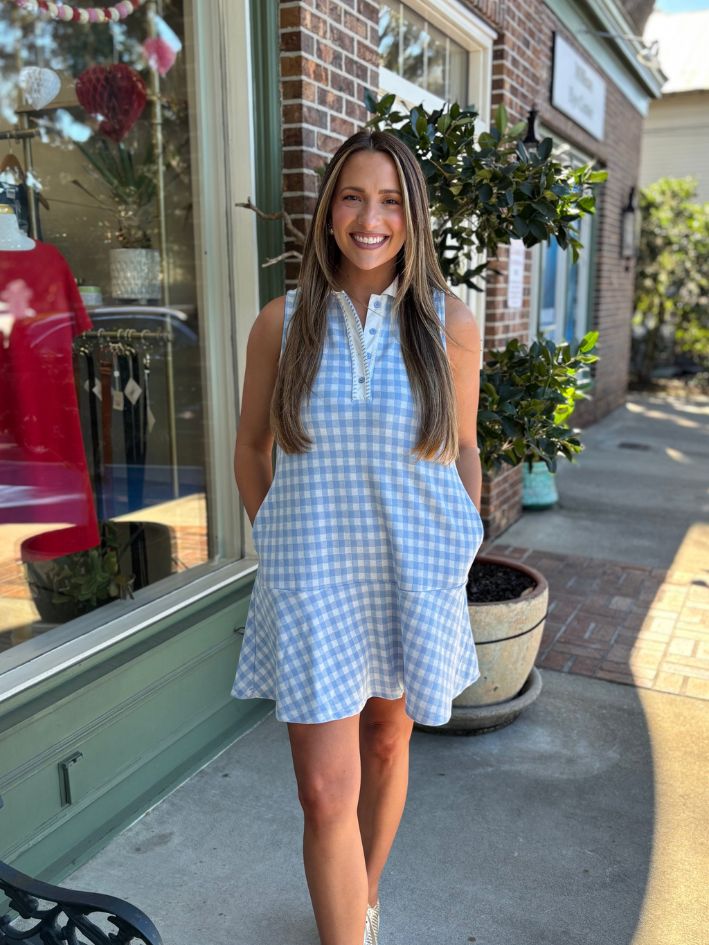 Woman in a blue checkered gingham dress standing on a sidewalk next to a store window.