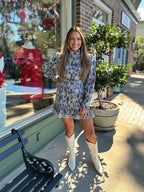 Woman in a patterned eyelet blue and white dress and white boots standing in front of a store window.