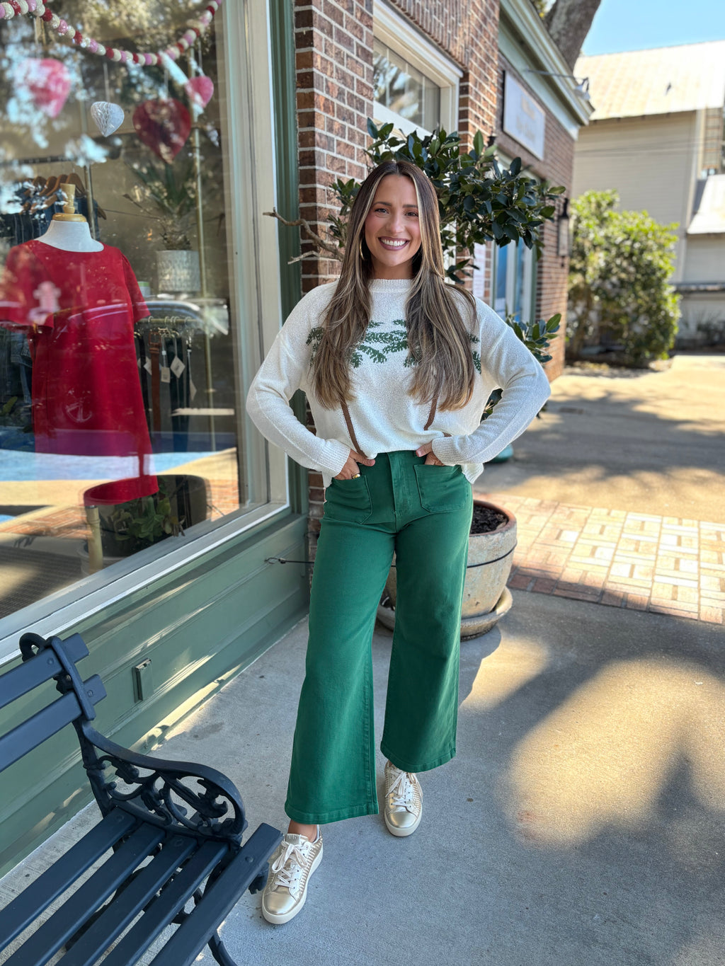 Woman wearing a white sweater with a green palm tree leaf pattern and green pants standing outside a store.