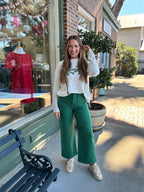 Woman wearing a white sweater with a green palm tree leaf pattern and green pants standing outside a store.