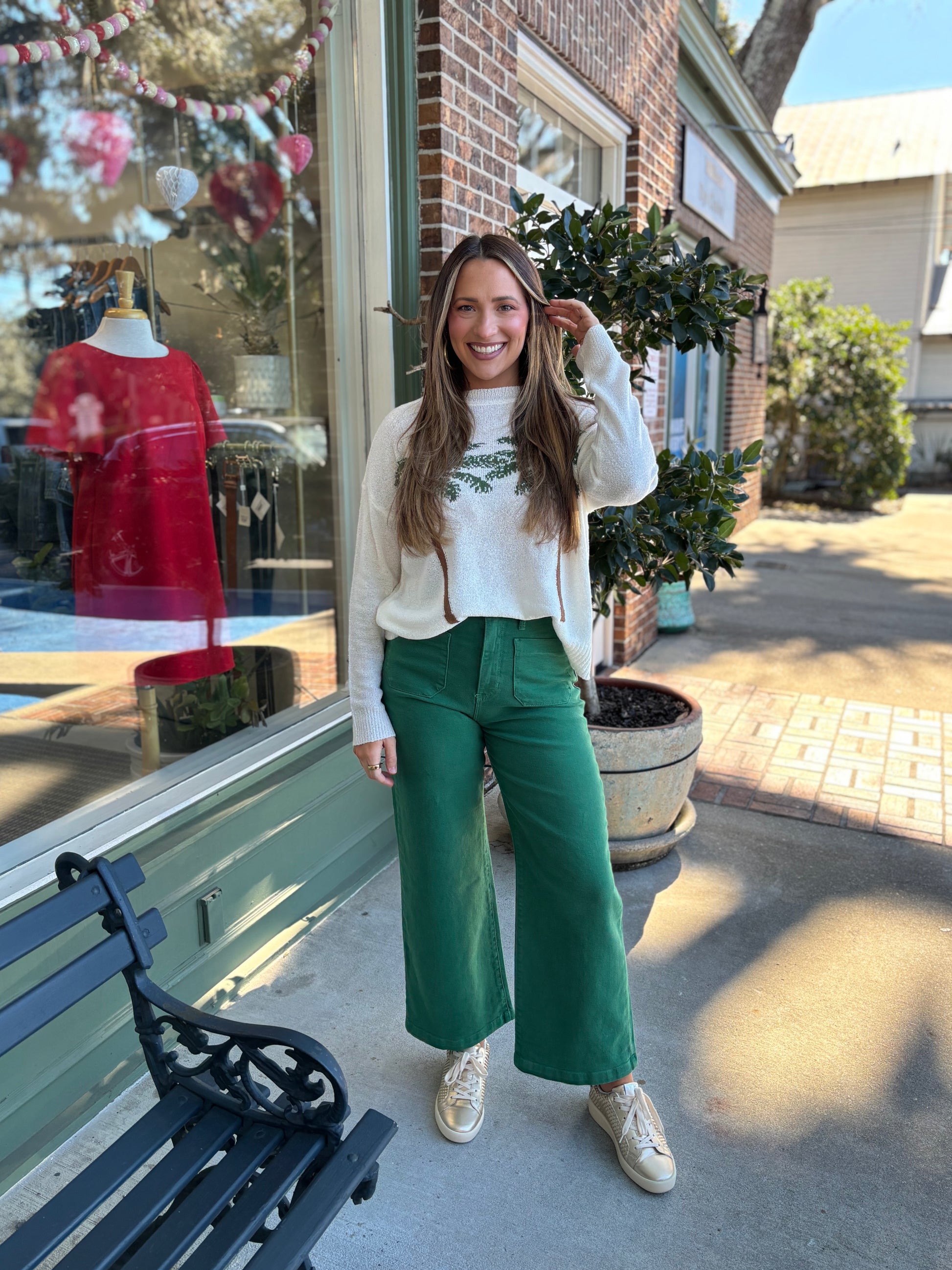 Woman wearing a white sweater with a green palm tree leaf pattern and green pants standing outside a store.