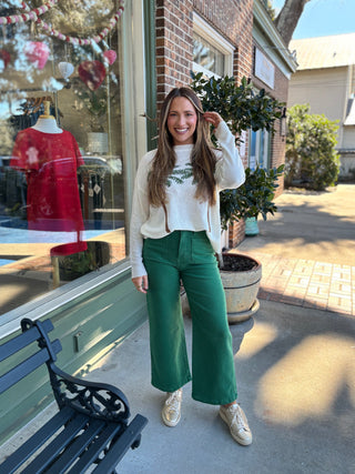 Woman wearing a white sweater with a green palm tree leaf pattern and green pants standing outside a store.