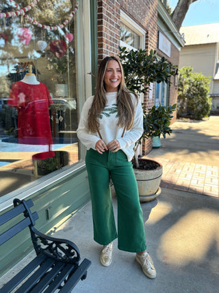 Woman wearing a white sweater with a green palm tree leaf pattern and green pants standing outside a store.